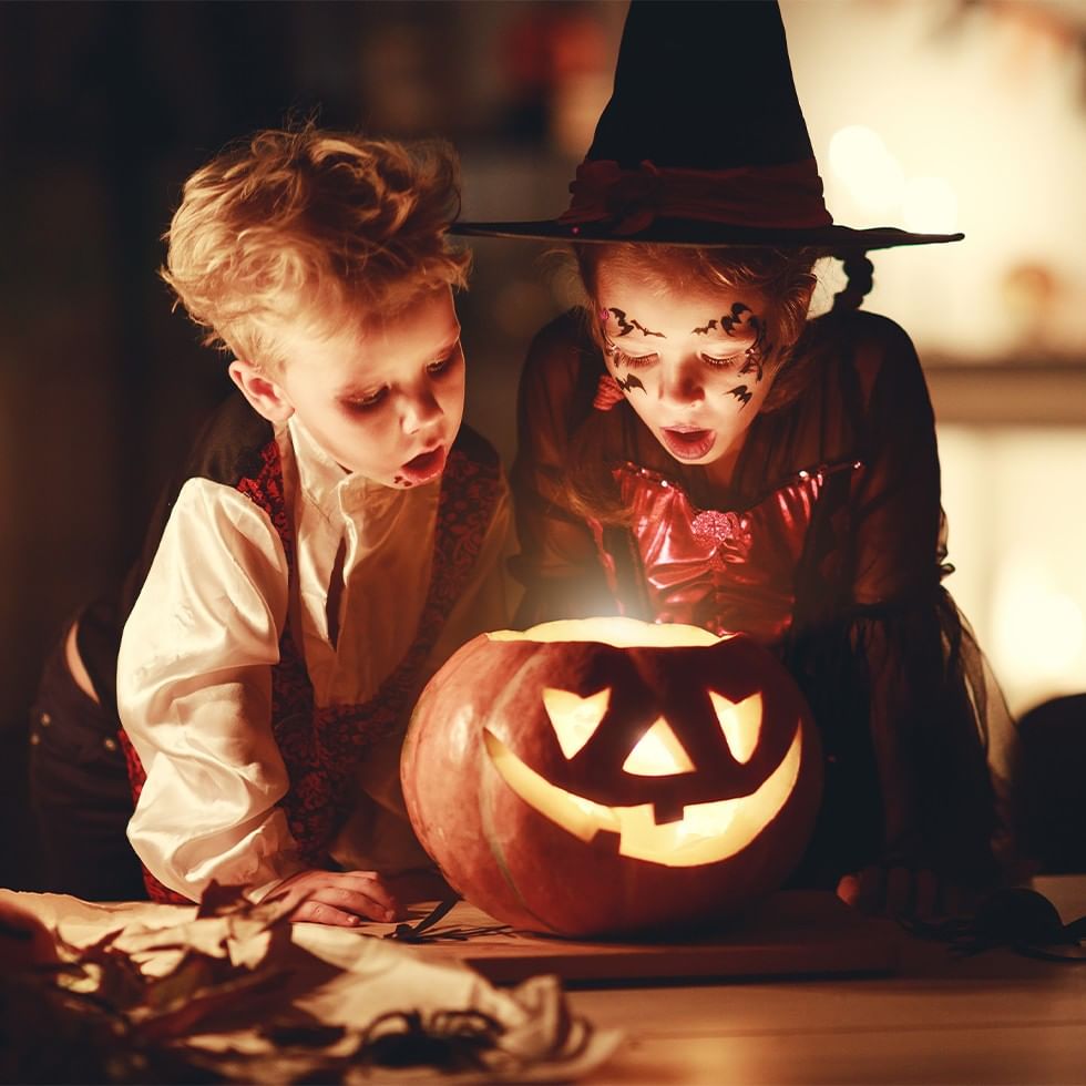 Zwei Kinder in Halloween-Kostümen betrachten einen Kürbis mit beleuchtetem Gesicht.