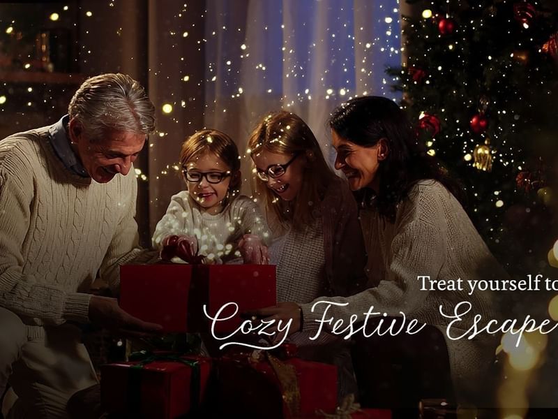 Family of four opening gifts near a Christmas tree with festive lights and text.