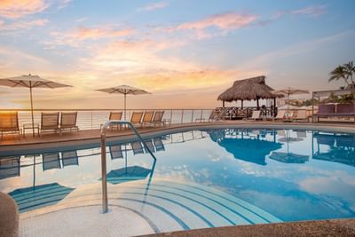 Piscina con tumbonas, sombrillas y un gazebo de techo de palma en el Sunset Plaza Beach Resort durante un atardecer con vistas a la playa de Puerto Vallarta