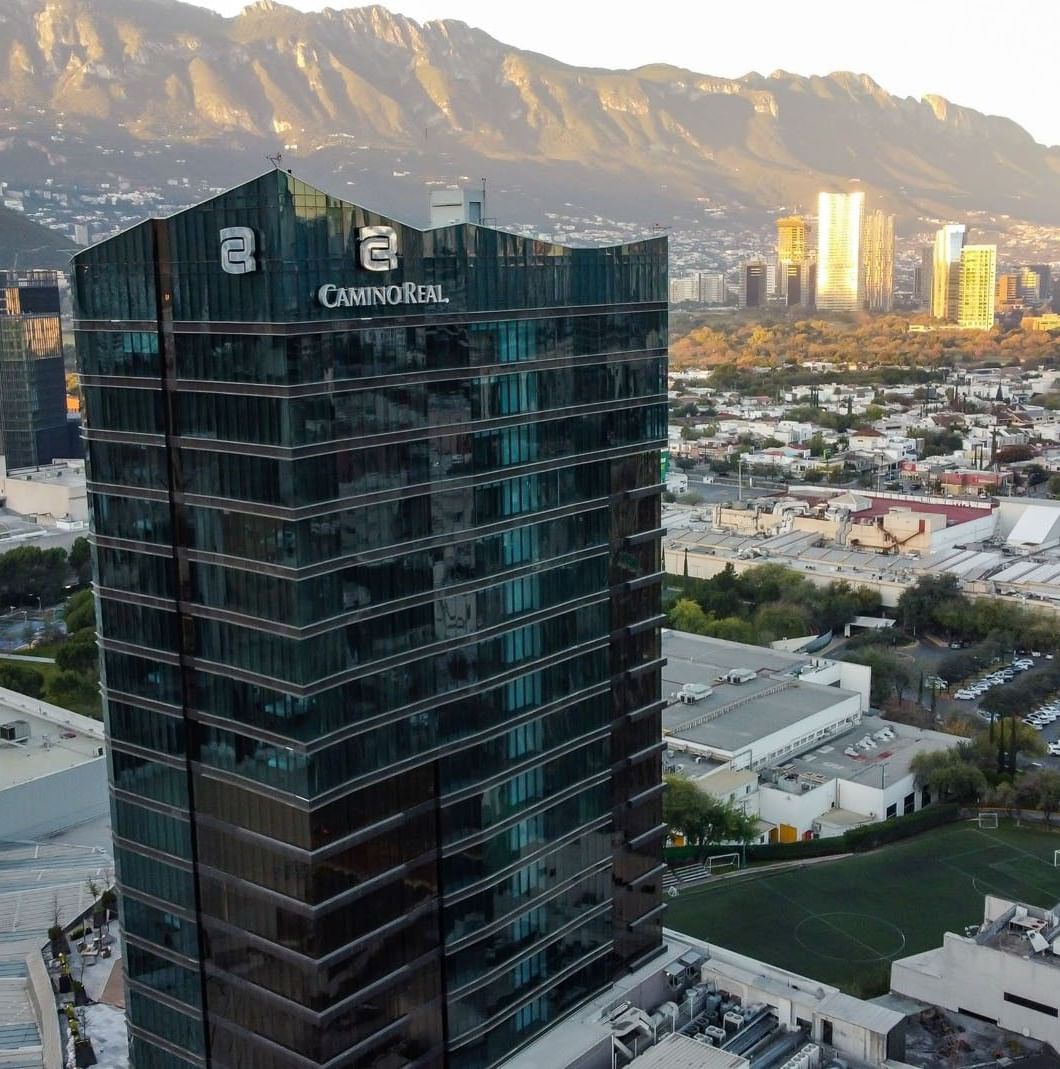 Aerial view of the Camino Real Fashion Drive Monterrey hotel, a glass skyscraper with mountains in the background