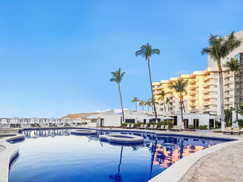 Large outdoor swimming pool at a luxury Sunvivia Mazatlán resort reflecting palm trees and white buildings under a blue sky