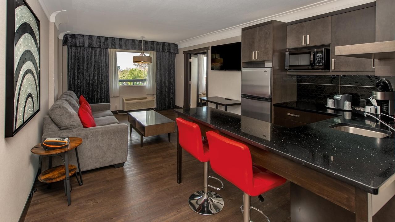 Modern suite with full kitchen, red bar stools, living area with gray sofa, and hardwood floors at Coast Calgary University Area Hotel by APA