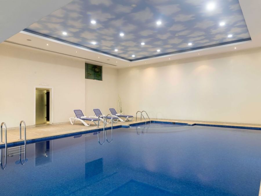 Indoor pool with blue tiles by striped lounge chairs under a cloud-patterned ceiling at Warwick Al Jubail