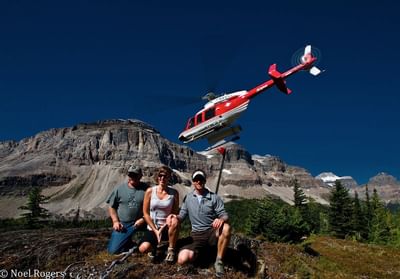 Helicopter soaring above a mountainous landscape while three people pose on rocky terrain near Blackstone Mountain Lodge