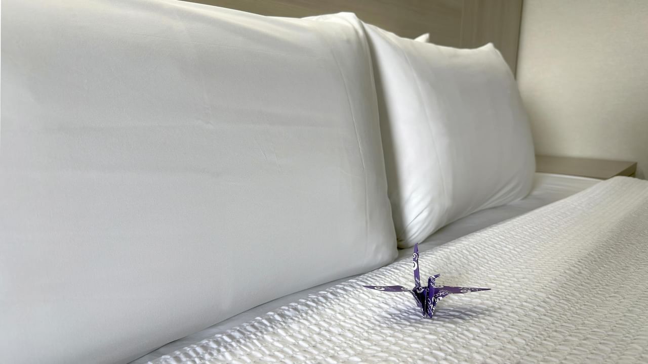 Neatly made bed with white pillows and textured bedding featuring a decorative purple origami crane accent.