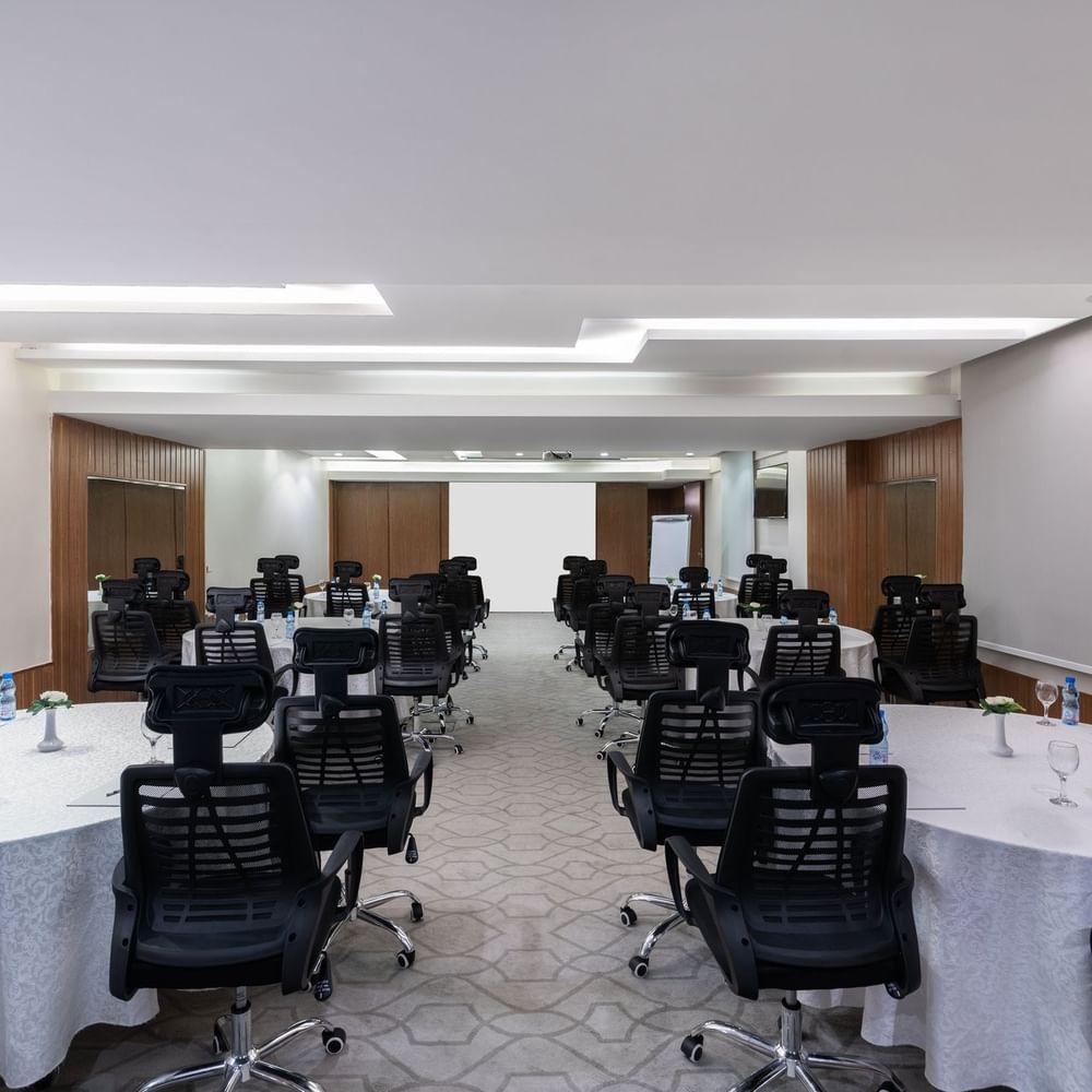 Round tables with rolling chairs facing a projector screen in Meeting Room B at Warwick Hotel Jeddah