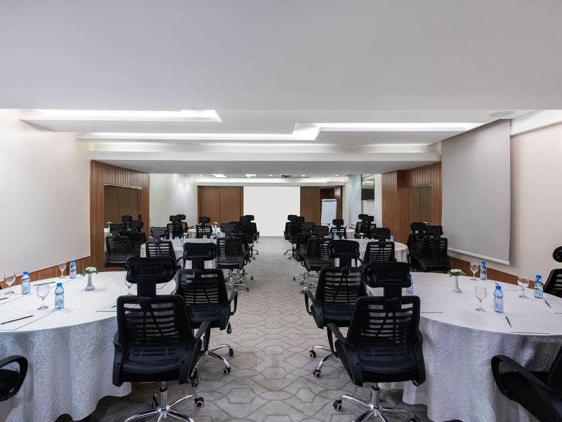Round tables with rolling chairs facing a projector screen in Meeting Room B at Warwick Hotel Jeddah