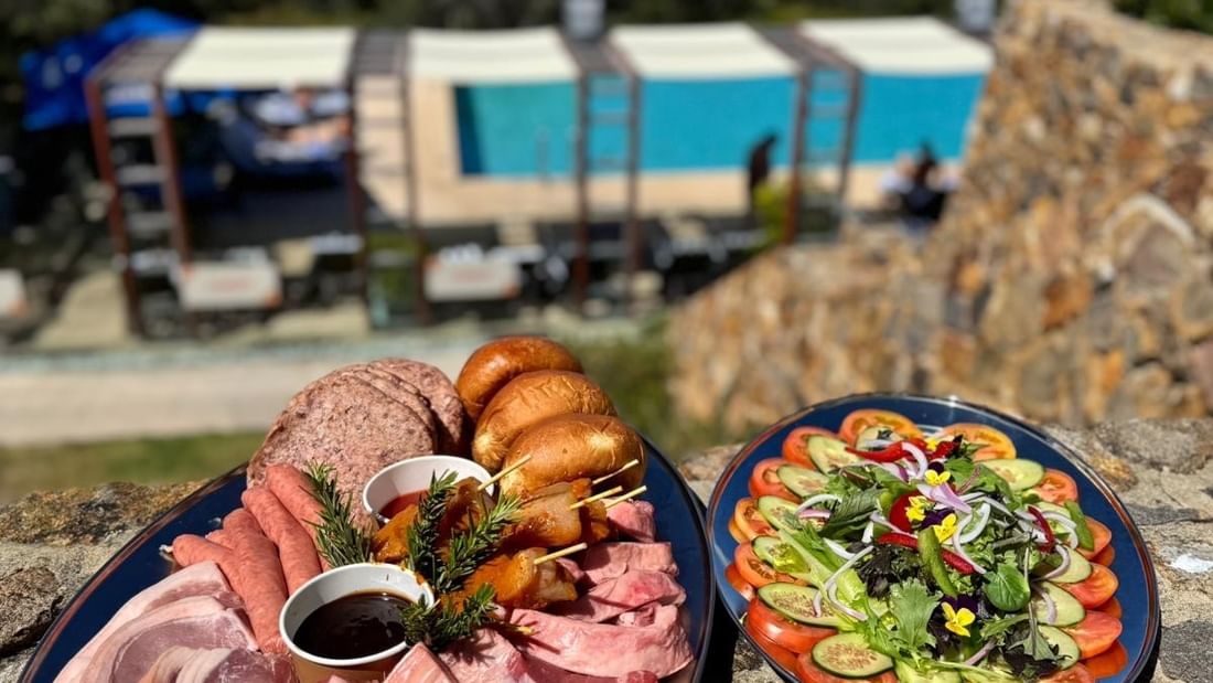 Plates of assorted meats, sausages, and salads on a stone surface with a pool view in the background.