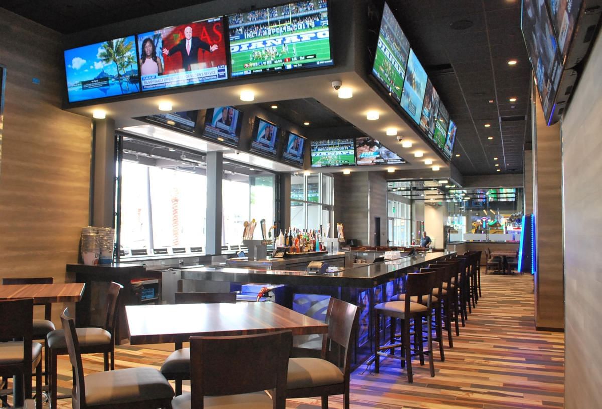 A bar with multiple screens showing sports, a counter with stools, and tables with chairs.
