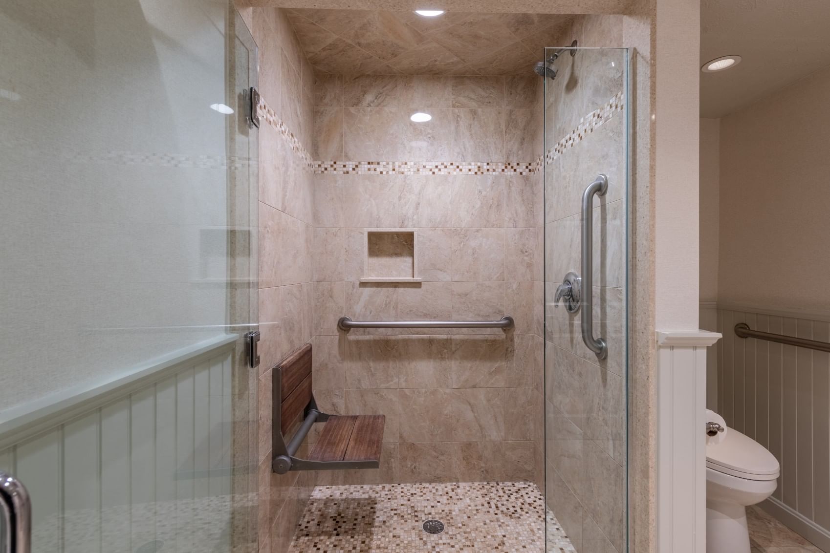 A bathroom shower with a glass door, grab bars, and a shower bench.