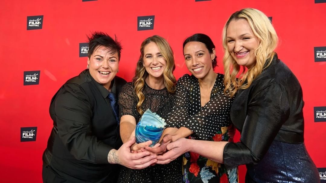 Four women smiling and holding a glass award in front of a red background with film festival logos.