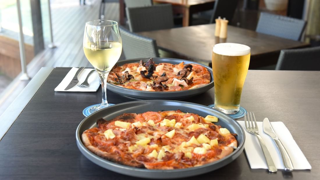 Two pizzas on plates, beside a glass of wine and a pint of beer in Celsius Restaurant & Bar at Mercure Hotel Townsville