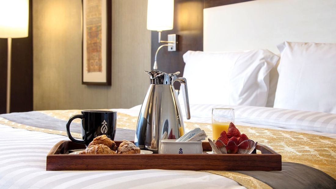 Breakfast tray on a bed with a silver coffee pot, orange juice, & croissants in the Deluxe Room at Warwick Hotel Jeddah