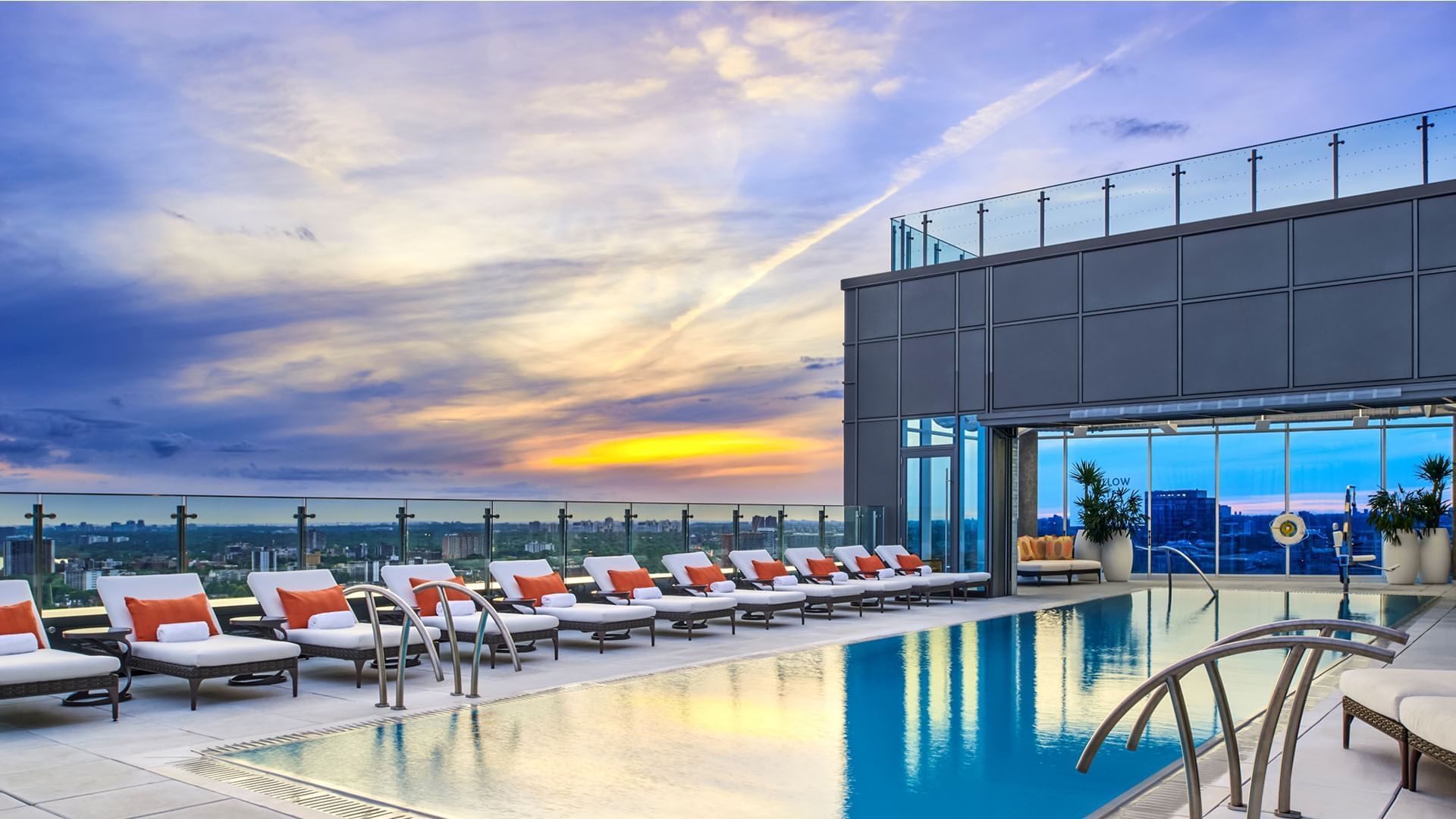 Outdoor rooftop pool with white lounge chairs and cushions facing colorful sunset city skyline at Hotel X Toronto