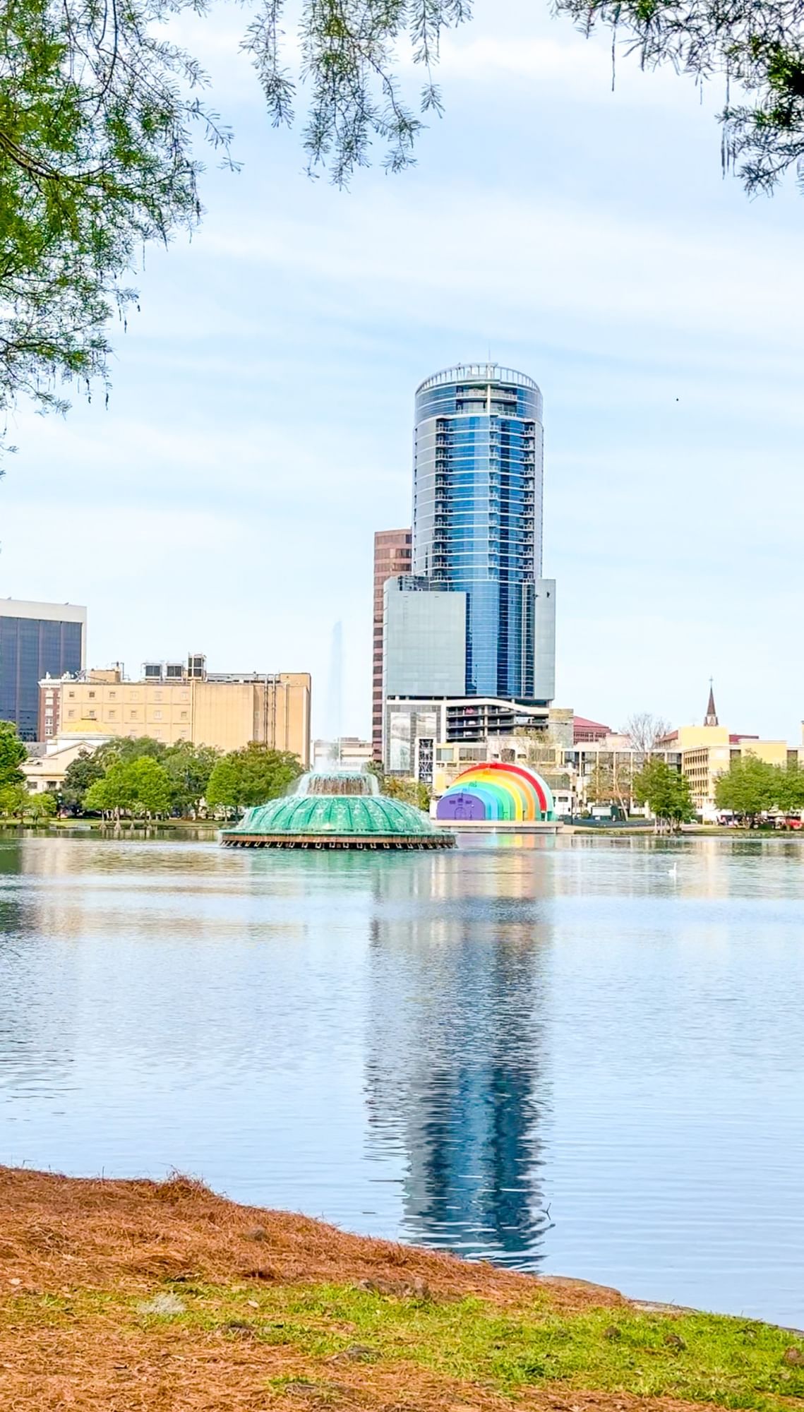 A large lake with a fountain in the middle surrounded by a tall building and several shorter buildings along with a rainbow painted outdoor theater. Lake Eola is a great budget-friendly activity to do for $25 or less in Orlando.