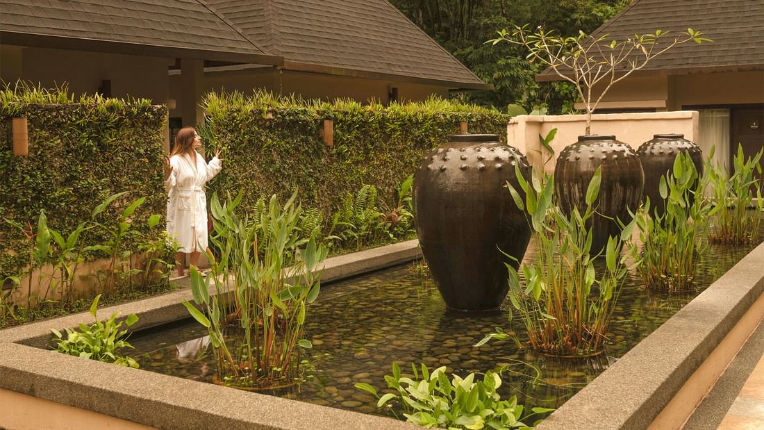 Woman in white robe walking near a small pond with lush greenery of The Banjaran Spa Centre