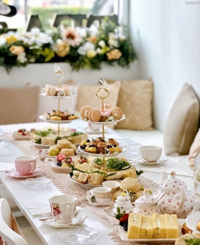 An elegant table setting with vintage floral teacups, tiered trays with pastries, tea pots, and plates of other treats. Beige pillows and a window box of flowers are out of focus in the background.