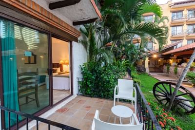 Balcony with white chairs and table, lush greenery, and a view of a yellow building with balconies.