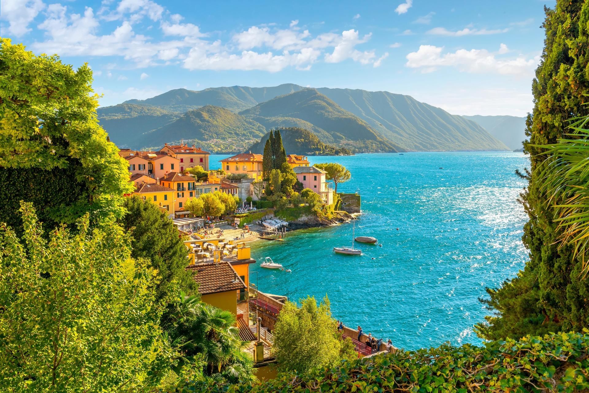 Scenic view of Lake Como with colorful buildings, lush greenery, and mountains under a bright blue sky near AG Hotels