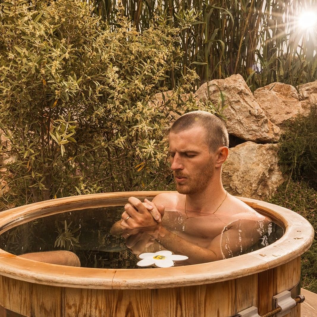 Hombre en un barril de madera de agua caliente rodeado de naturaleza en Luminaries in Bloom David Seed.