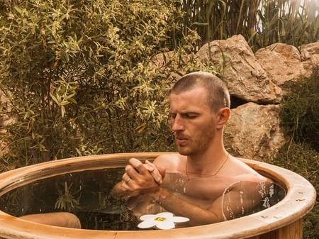 Man relaxing in wooden hot tub with a flower floating in water.
