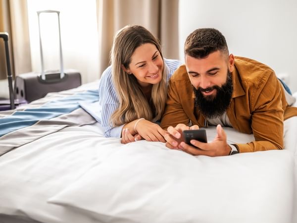 Couple lying on bed, looking at smartphone, suitcases nearby.