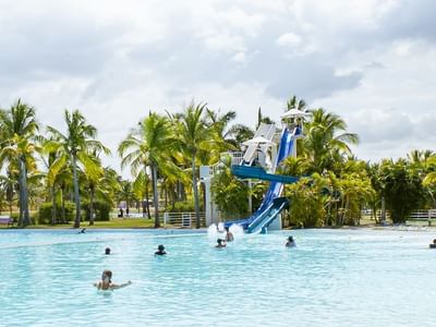 Large turquoise pool with water slide and palm trees under cloudy sky at Playa Blanca Beach Resort
