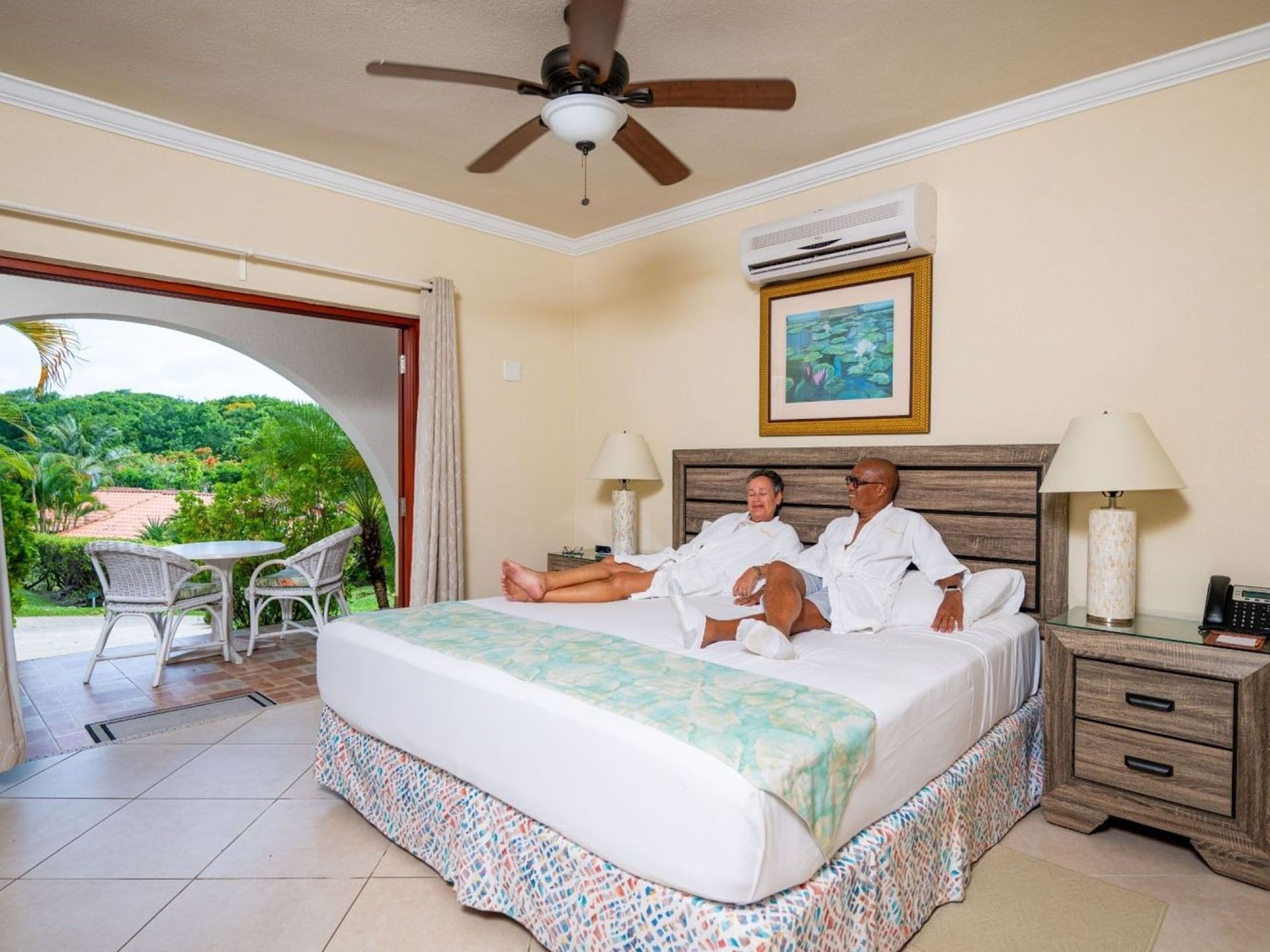Couple relaxing on a king bed beside an open terrace door with a view of lush trees at Sugar Cane Club Hotel & Spa