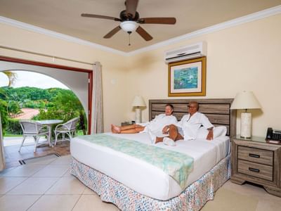 Couple relaxing on a king bed beside an open terrace door with a view of lush trees at Sugar Cane Club Hotel & Spa