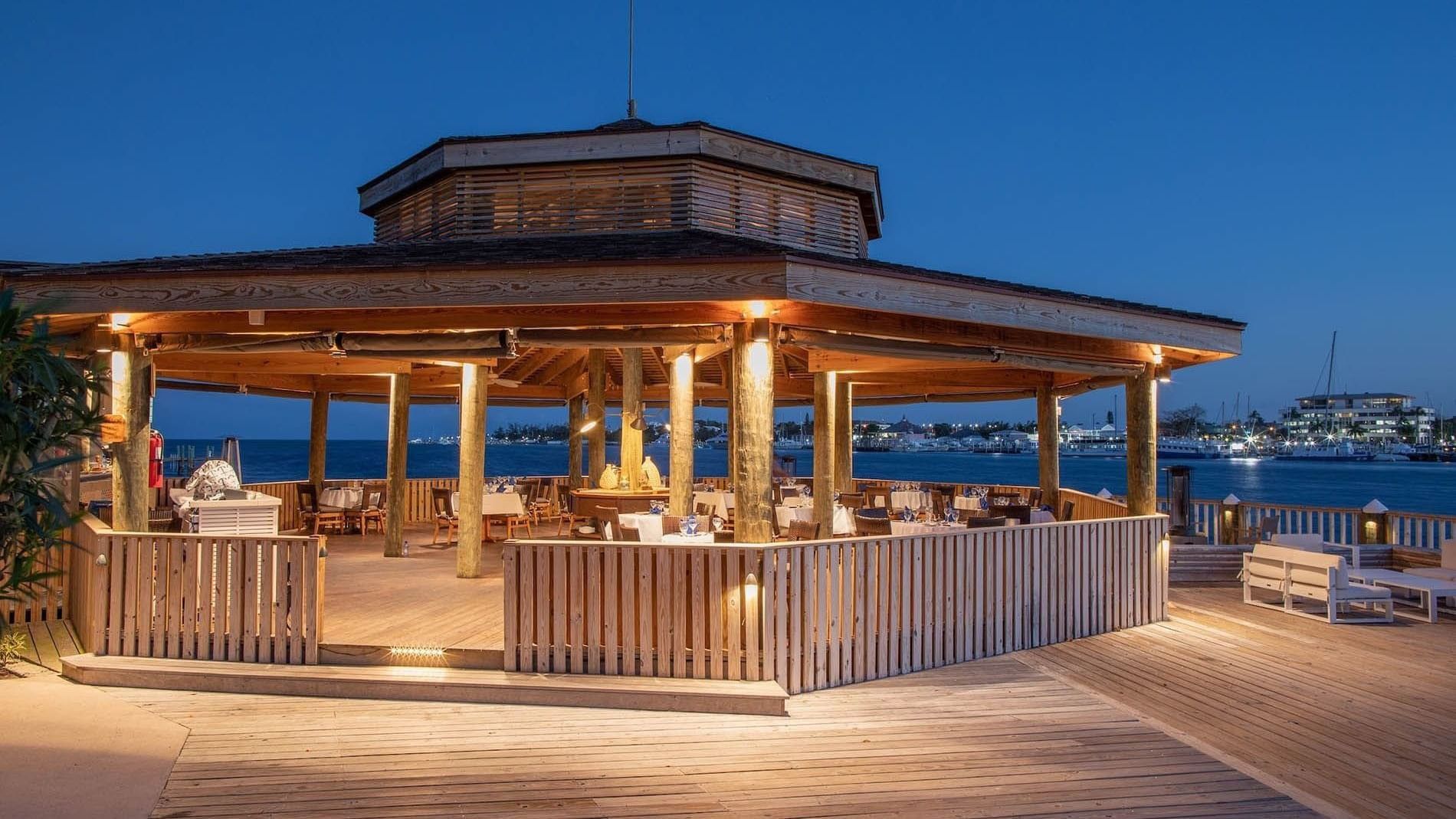 Circular bar by dining tables under a wooden roof surrounding a night harbor at Warwick Paradise Island Bahamas