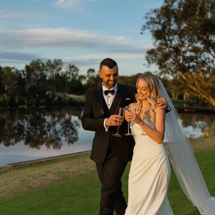 A couple in wedding attire toasting with champagne by a lakeside near Mercure Kooindah Waters