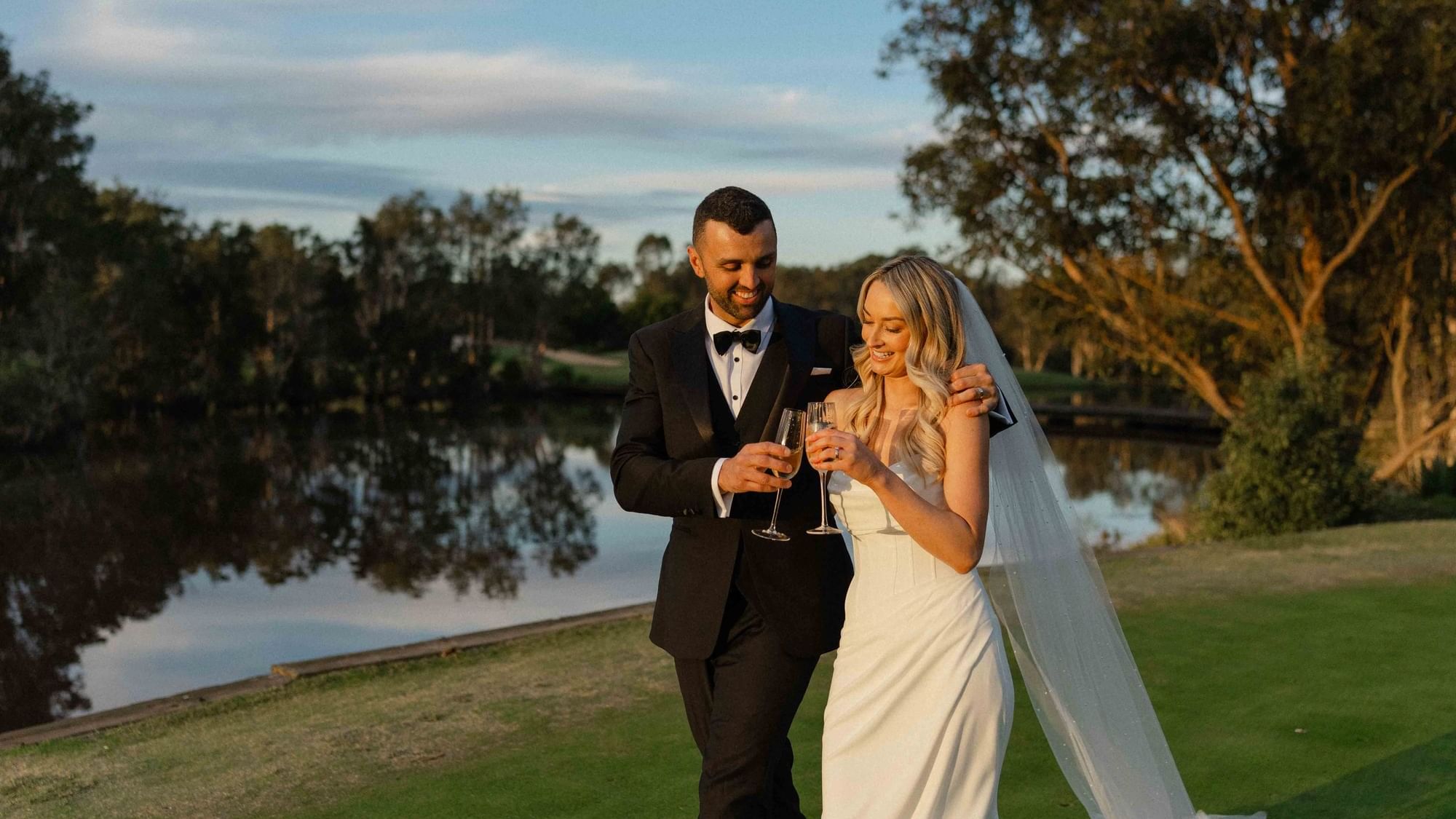 A couple in wedding attire toasting with champagne by a lakeside near Mercure Kooindah Waters