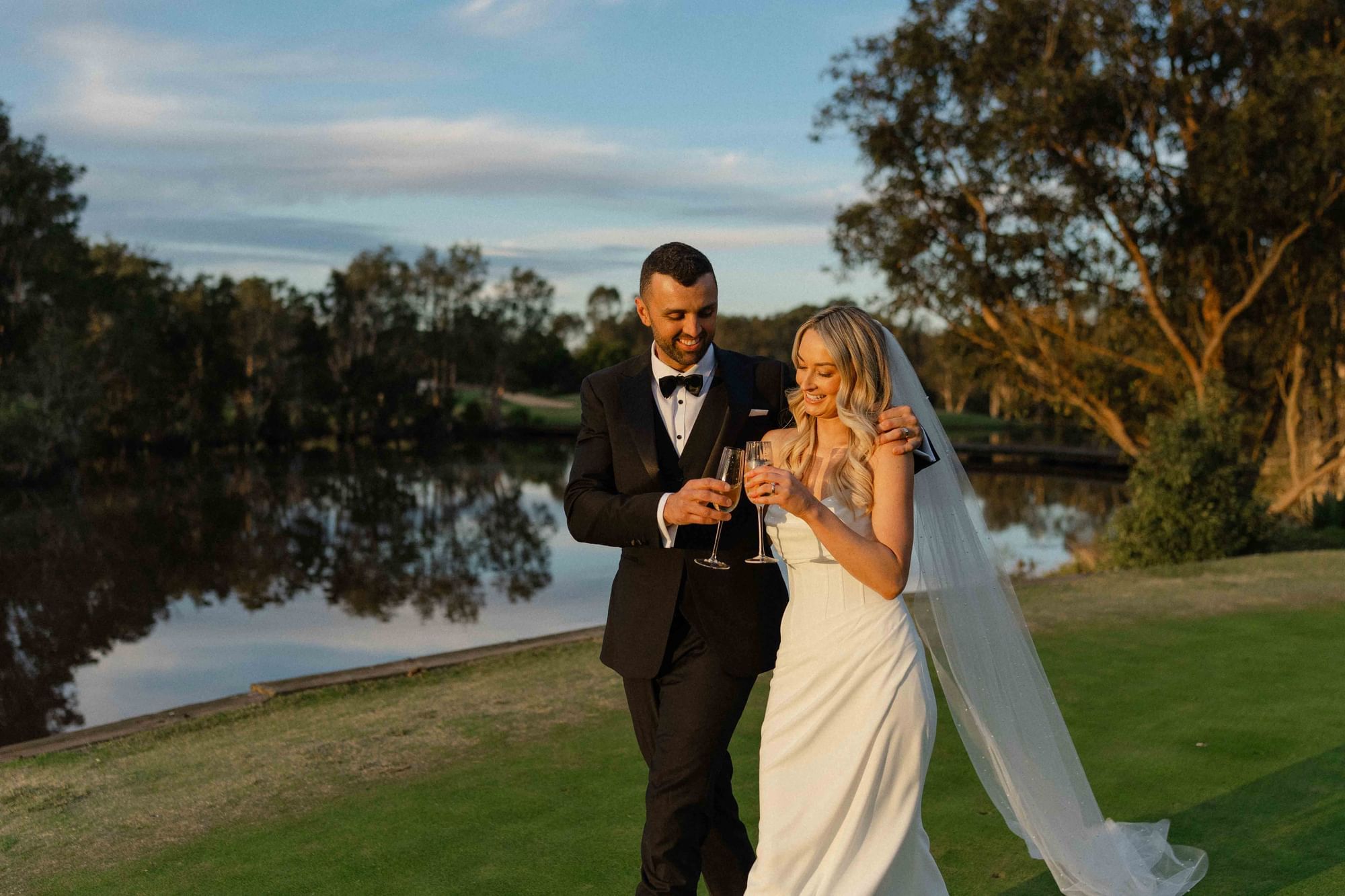 Bride and groom holding champagne glasses by a lake near Mercure Kooindah Waters