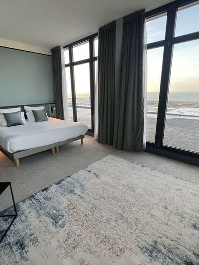 Chambre d'hôtel avec lit, tapis, fenêtres panoramiques et vue sur la mer.