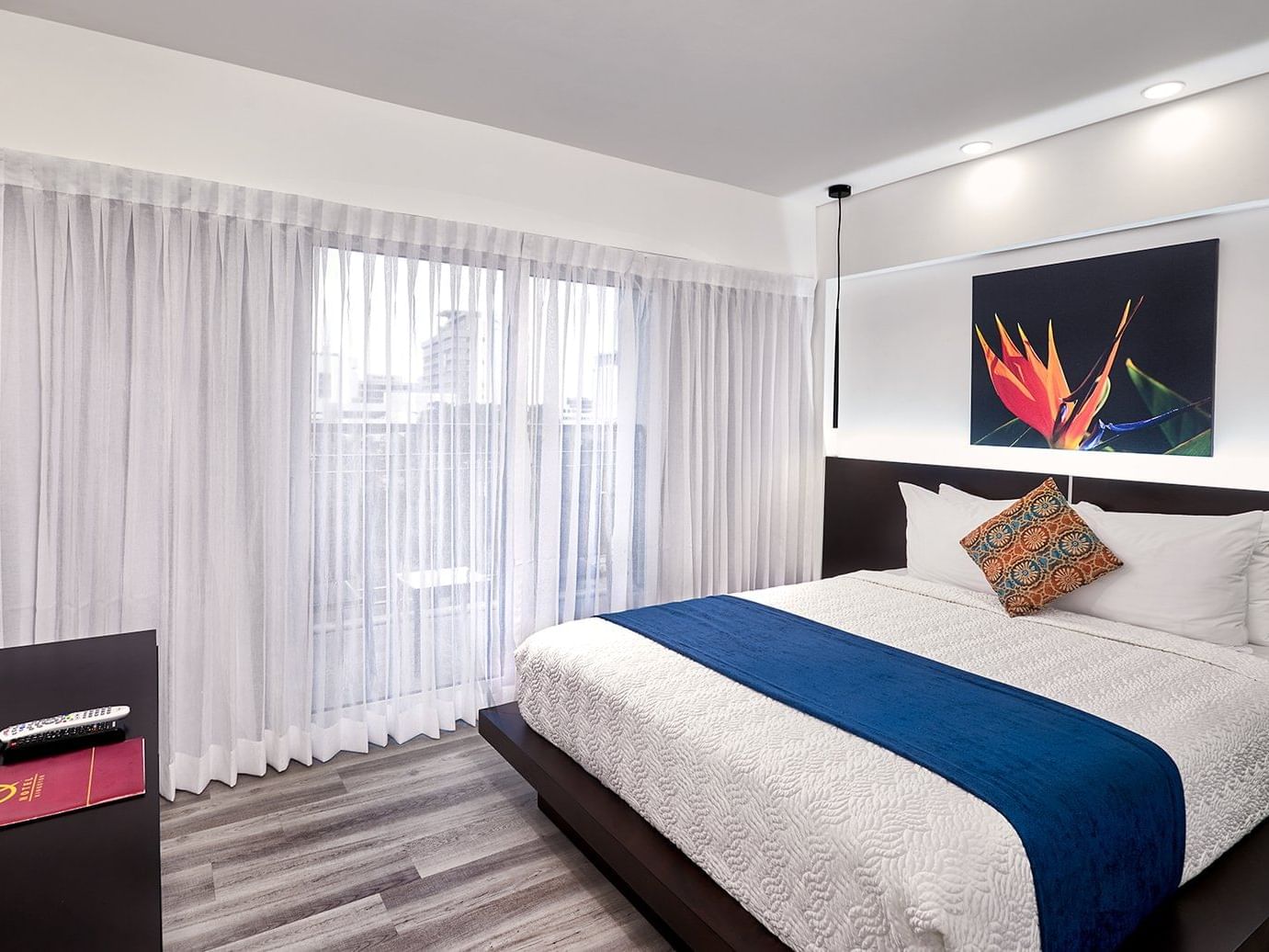 Modern bedroom with platform bed, blue runner, white curtains, and vibrant floral artwork at R Hotel Kingston.