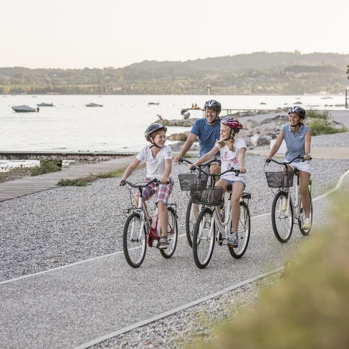 Family of four riding bicycles on a gravel path by the waterfront, wearing helmets and smiling.