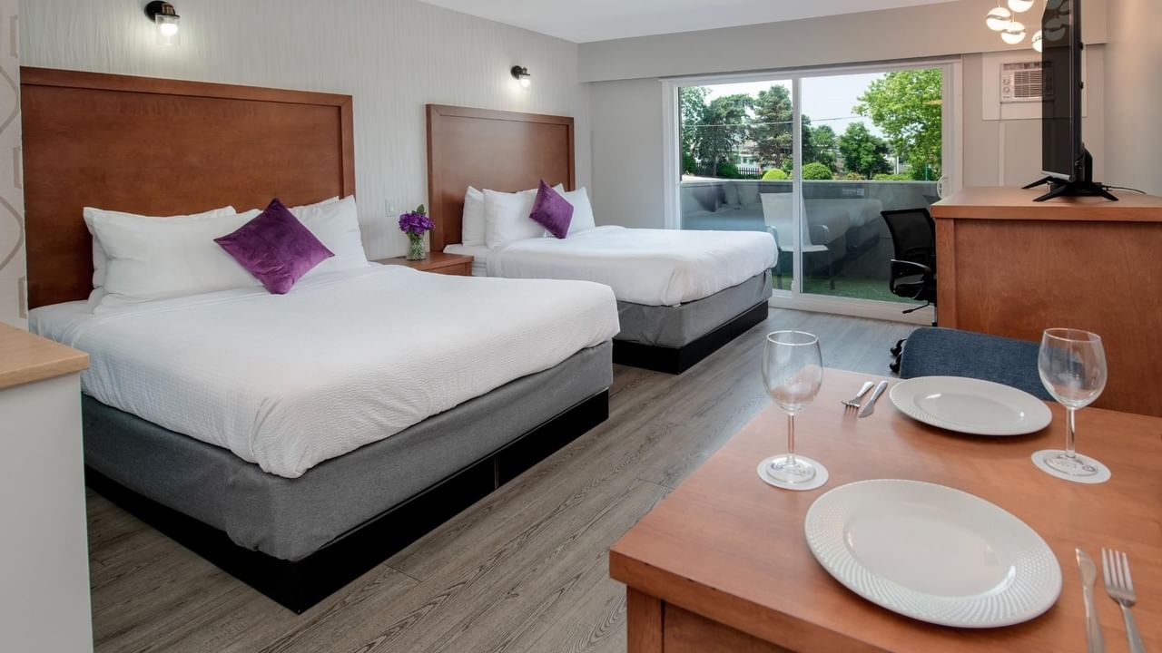 Two queen beds with white linens and purple pillows, a dining table with plates and wine glasses, and a balcony view.