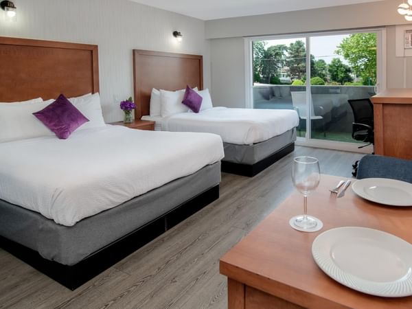 Two queen hotel beds with purple pillows, a dining table and a balcony with a view.