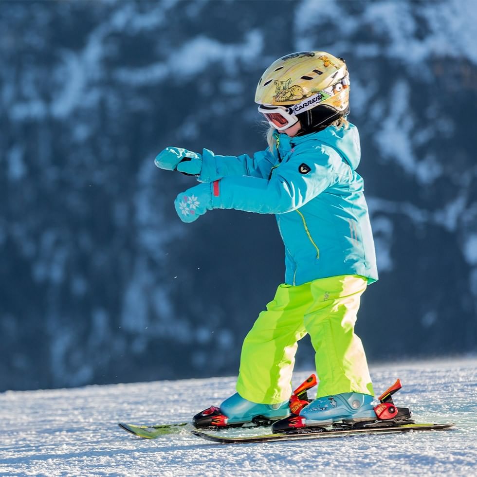 Kind im Skianzug und Helm steht auf Skiern im Schnee vor verschneiten Bergen.