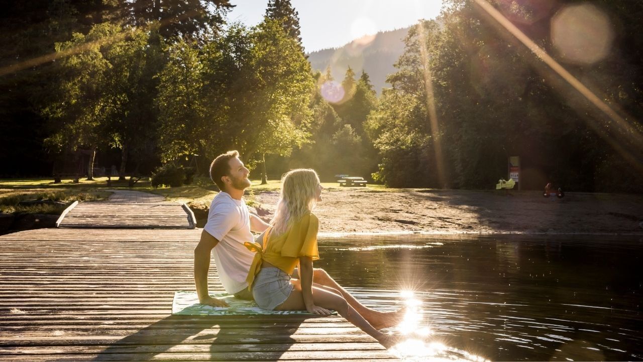Two people sitting on a wooden dock with feet in the water, surrounded by trees and mountains in golden sunlight.