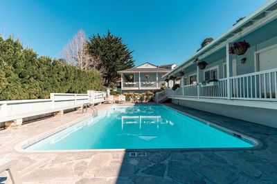 The sunny outdoor swimming pool surrounded by a stone patio and the light blue hotel buildings at Carmel Bay View Inn