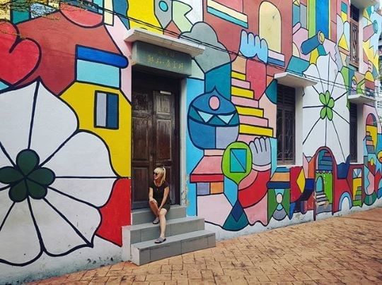 A woman sits on steps beside a vibrant, geometric mural featuring bold colors and playful shapes near YTL Hotels