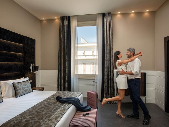 Couple dancing joyfully by the bed with large windows in Superior Room at The Glam Hotel