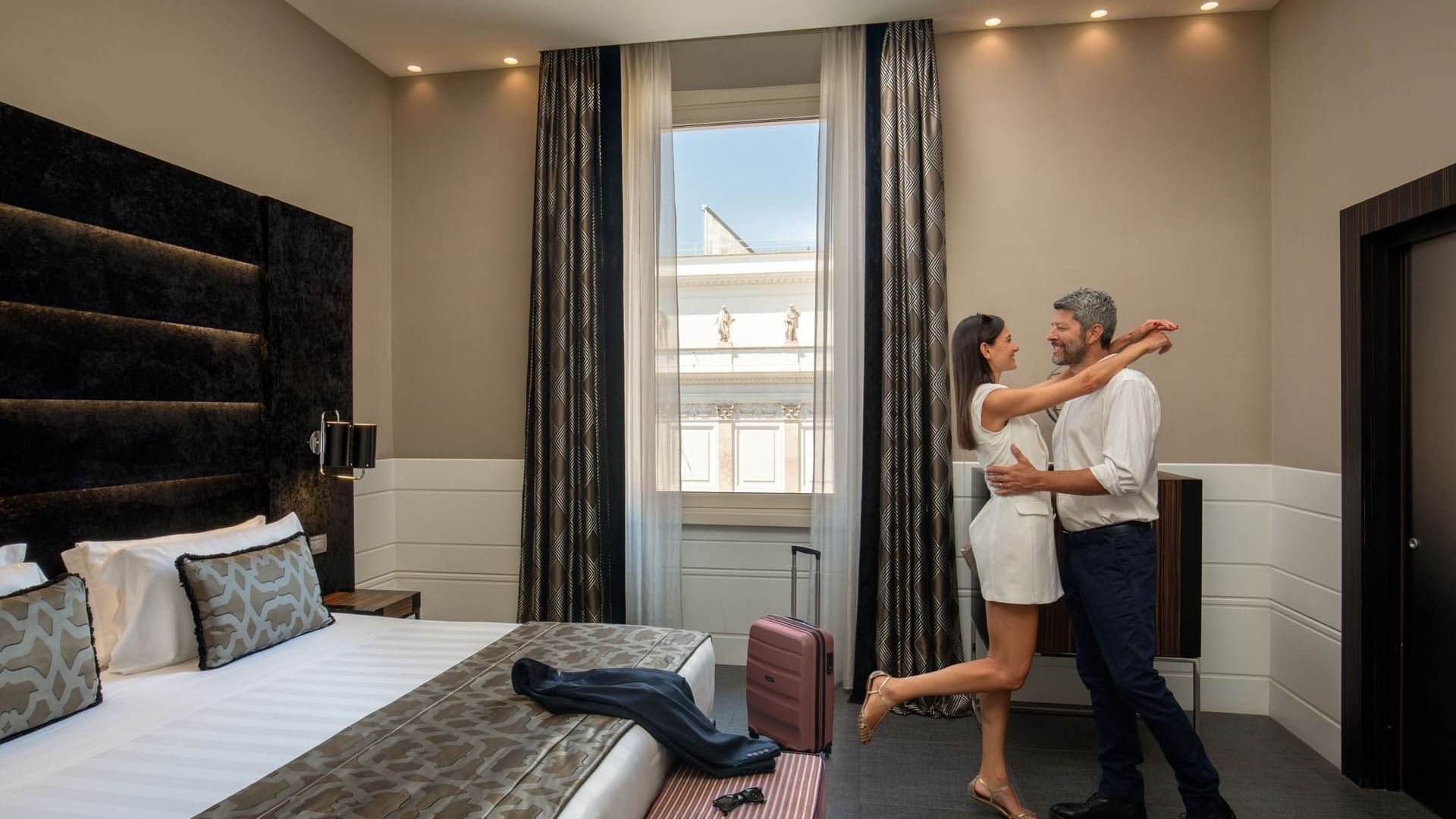 Couple dancing joyfully by the bed with large windows in Superior Room at The Glam Hotel