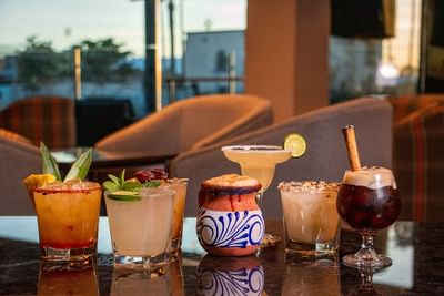 Vibrant cocktails with elegant garnishes lined up on a dark counter by comfy lounge chairs in El Colorado Bar at Hotel Mirra