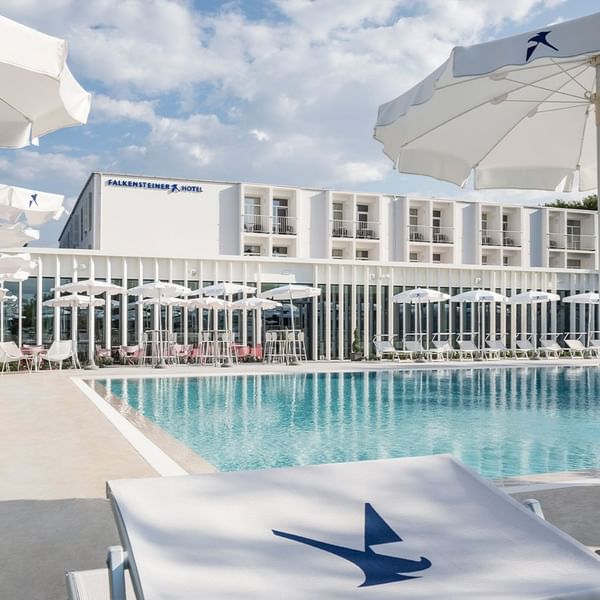 Hotel Falkensteiner mit Pool, Liegen, Schirmen und modernem Gebäude im Hintergrund unter blauem Himmel.