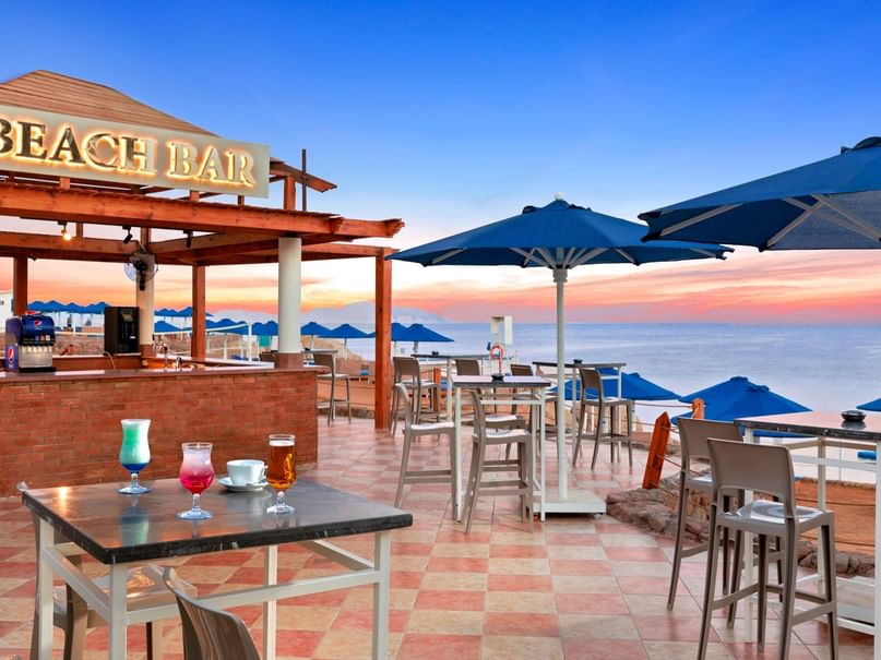 Beach Bar sign above an outdoor bar with tables, chairs, and drinks, overlooking the ocean at sunset.