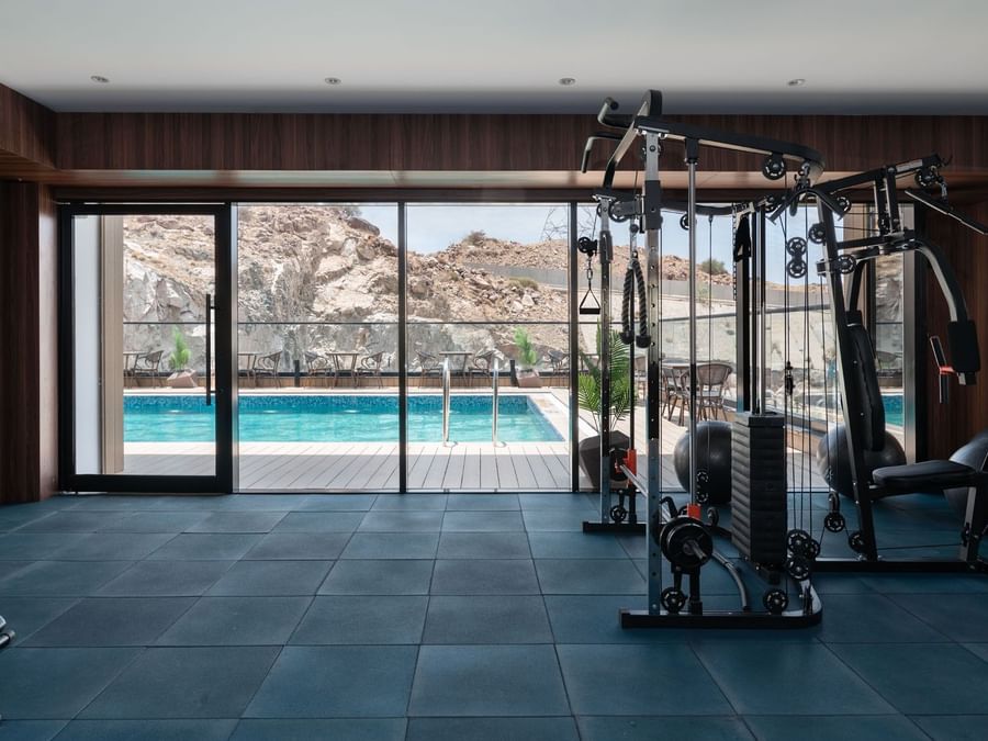 Gym equipment by a pool under a clear sky seen through large windows at Warwick Al Taif Hotel