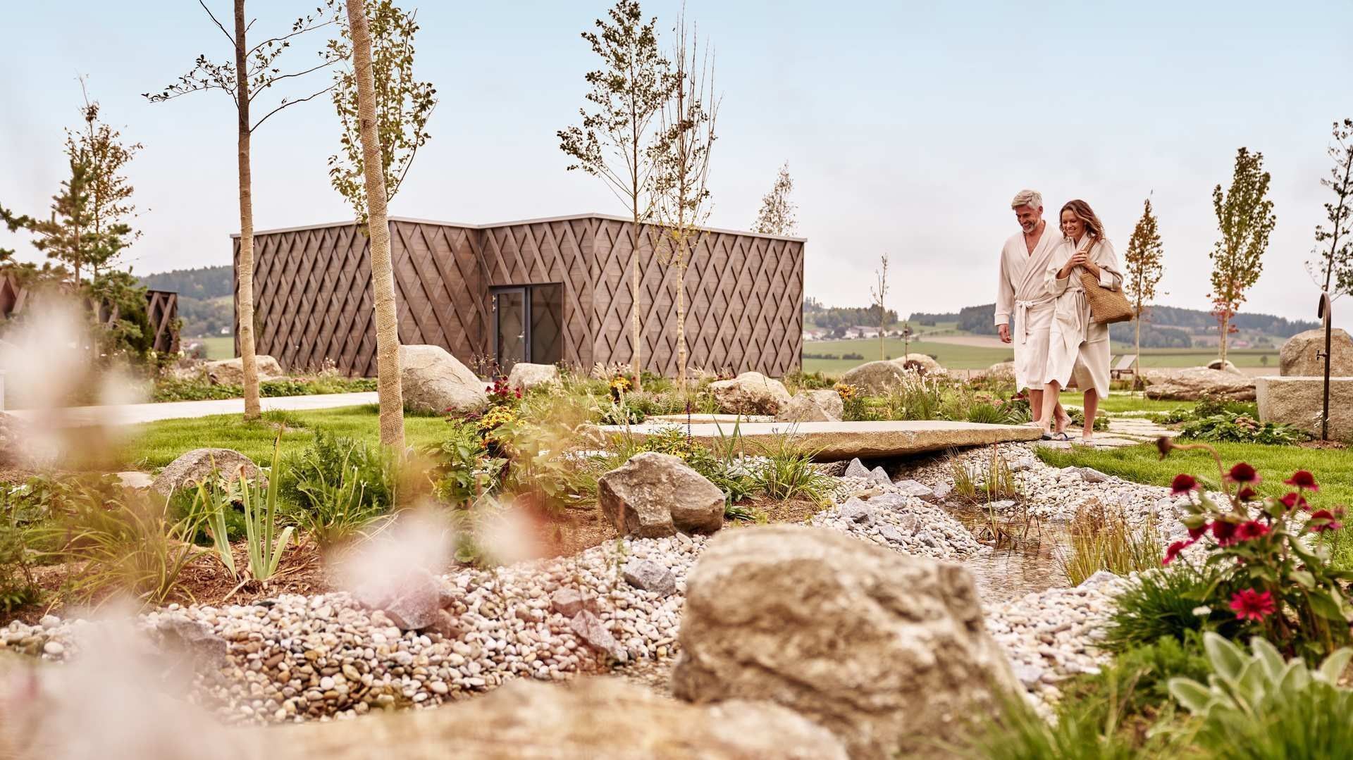 Coppia in abiti da bagno attraversa un ponte di pietra in un giardino con rocce, piante e un edificio.