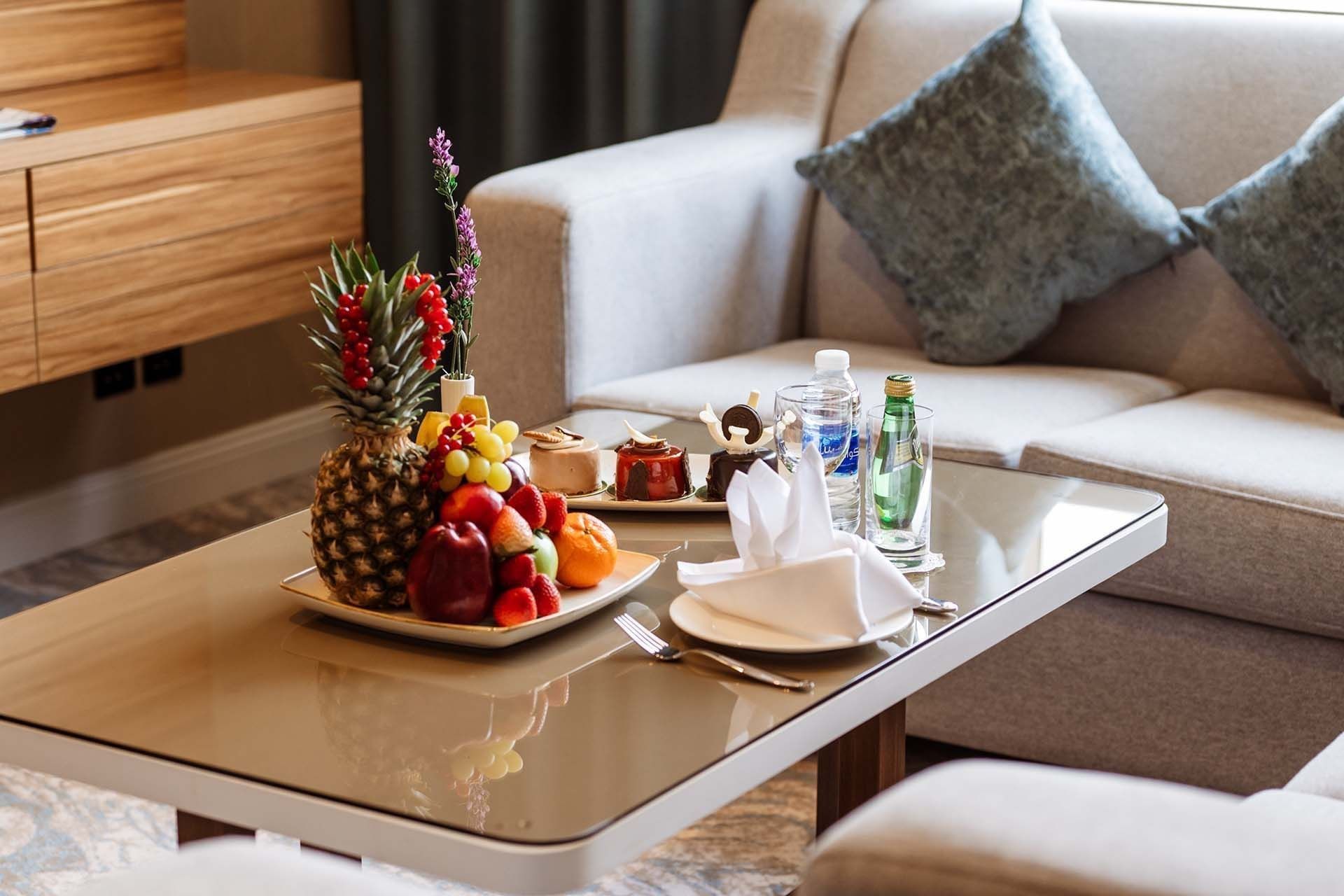 Fruit platter and pastries on a glass table by a grey sofa in 24/7 In Room Dining at Cantonal Hotel by Warwick Riyadh